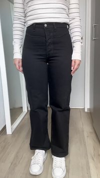 Jeans acampanados de mezclilla negra con una cintura alta, diseño de cinco bolsillos y costuras claras, combinados con zapatillas blancas sobre un suelo de madera.