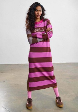 Kobieta o długich kręconych włosach stoi ze skrzyżowanymi ramionami, ubrana w różowo-brązową sukienkę w paski z napisem „Sugar & Spice” oraz kolorowymi naszywkami z kreskówkami.