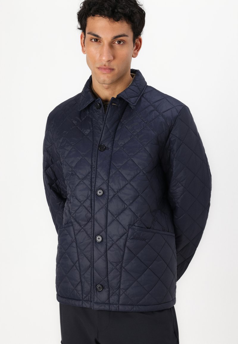 Veste matelassée marine avec un col, des boutons sur le devant, deux poches latérales et un motif en losanges cousu. Légère, texture lisse.