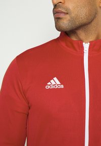 Czerwona sportowa kurtka z wysokim kołnierzem, białym zamkiem błyskawicznym i białym logo Adidas po lewej stronie na klatce piersiowej. Gładka tekstura i dopasowany krój.