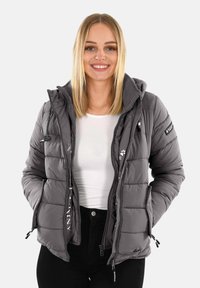 Woman smiling, wearing a gray quilted puffer jacket over a white shirt and black pants, hands in jacket pockets, against a plain background.