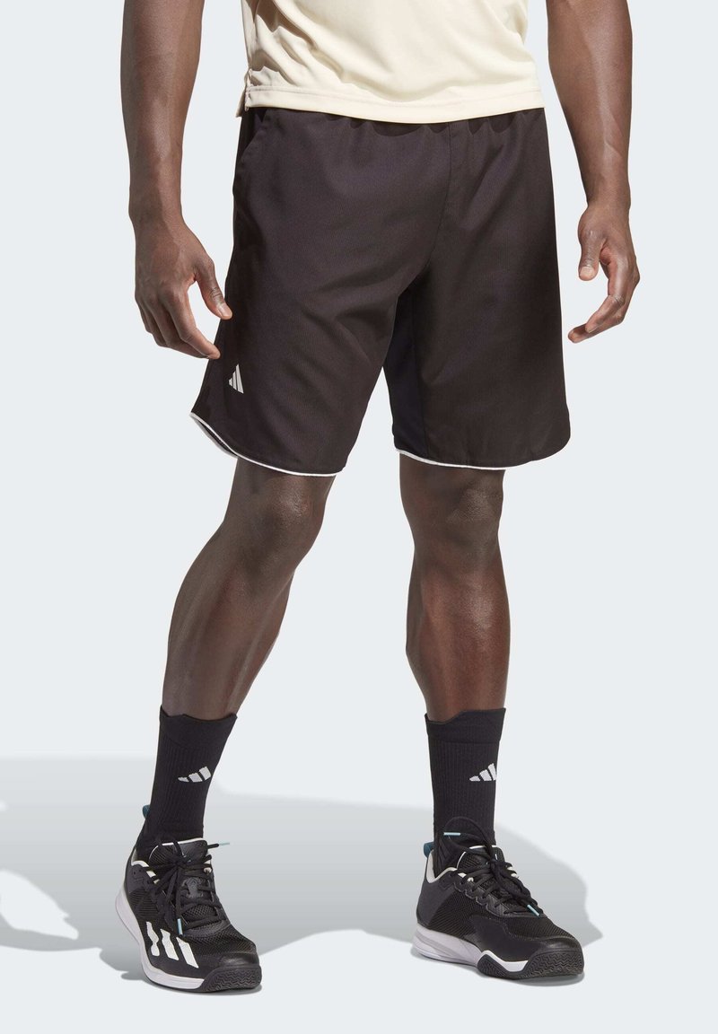 Juodos sportinės šortai su lygiu paviršiumi, balta apvadu palei kraštą, kairėje pusėje vaizduojamas Adidas logotipas. Modelis dėvi juodas kojines ir batelius.