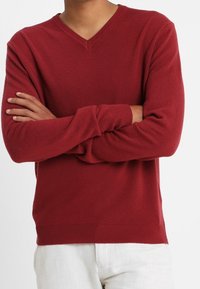 Person med korsade armar som bär en röd V-ringad tröja och vita byxor mot en enfärgad bakgrund.