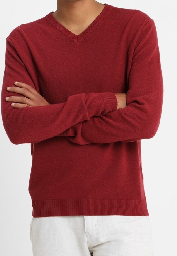 Person med korsade armar som bär en röd V-ringad tröja och vita byxor mot en enfärgad bakgrund.