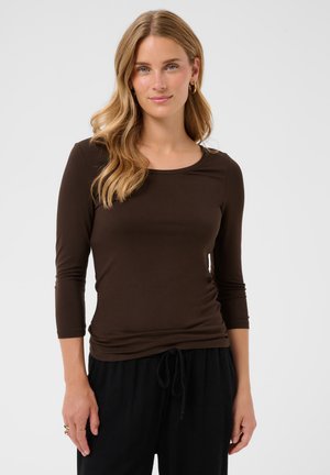 Donna bionda con capelli mossi sciolti che indossa una maglia aderente marrone scuro a maniche lunghe e pantaloni neri con coulisse su sfondo grigio.