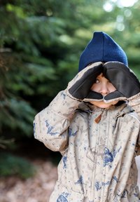 Beige patterned jacket with blue nature motifs, navy hat, and black mittens. Child makes binocular shape with hands, standing in a forest.