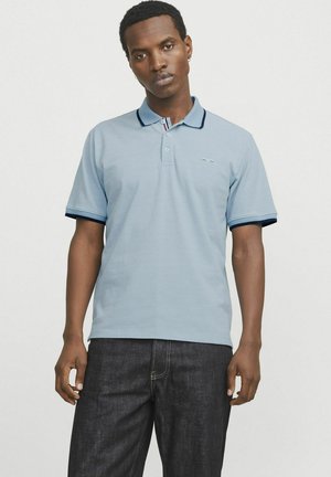 Lysblå polo med krave, med marineblå accenter på ærmerne og et lille logo på brystet. Kombineret med mørke jeans.