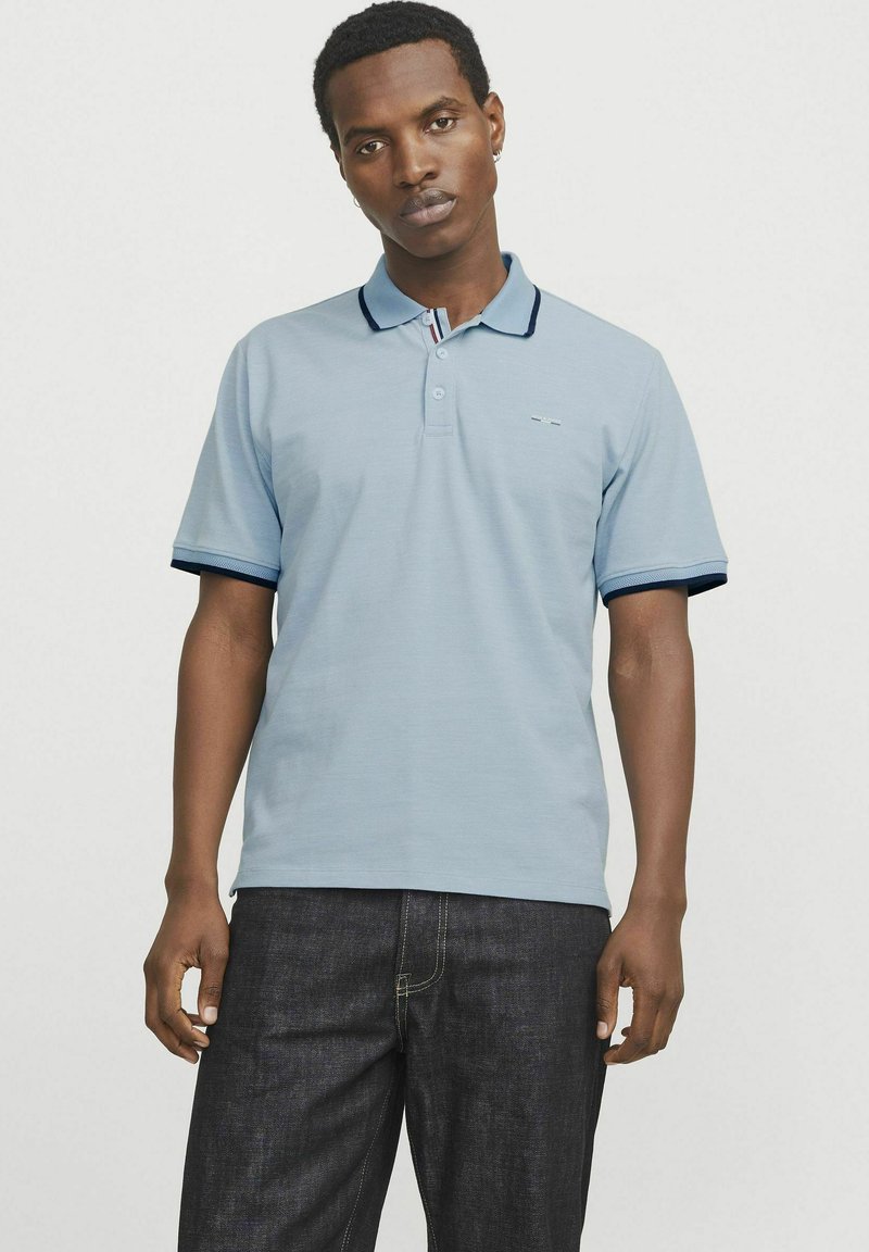 Ljusblå polo med krage, med marinblå detaljer på ärmarna och en liten logotyp på bröstet. Matchas med mörka jeans.