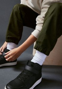 Personne portant un pantalon de jogging vert olive et des chaussettes blanches, en train de fermer des baskets noires montantes avec des lacets et une bride velcro, assise.