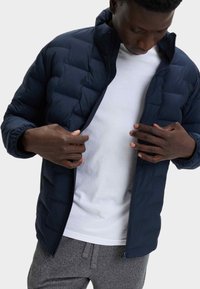 Chaqueta de plumas azul marino con cuello alzado y cremallera en el frente, con paneles acolchados y puños elásticos, llevada sobre una camiseta blanca.