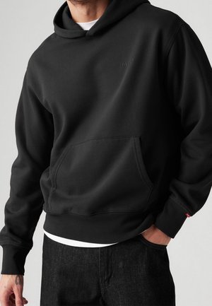 Mand iført en sort Levi's hættetrøje med frontlomme, kombineret med mørke jeans og en hvid undertrøje synlig ved kanten.