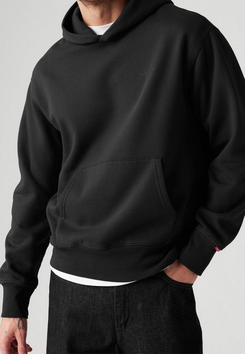 Homme portant un sweat à capuche noir Levi's avec poche avant, associé à un jean foncé et un sous-vêtement blanc visible à l'ourlet.