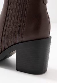 Brązowy skórzany botek za kostkę z pionowym przeszyciem i masywnym czarnym obcasem, o gładkiej fakturze i zaokrąglonym kształcie.