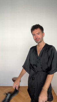 Črni satenast plašč z kratkimi rokavi, pasom s kravato in gladko teksturo. Ozadje je nevtralno. Roka drži črno dodatno opremo.
