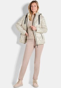 Giacca trapuntata di un leggero beige con lacci neri e cerniere, indossata sopra un maglione a maglia beige e pantaloni rosa chiaro, abbinata a stivali beige.
