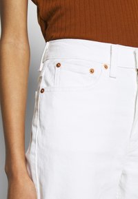 Nederdel av person som bär vita denimjeans och en brun ribbad ärmlös topp, som visar midjan, fickknappar och arm.