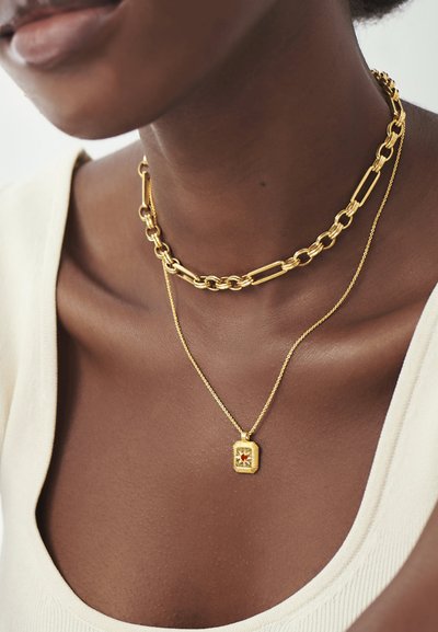 Guldtonet lagdelt smykker med en kraftig kæde-choker og en fin kæde-halskæde med en rektangulær vedhæng og en central rød ædelstenskant.