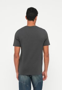 Homem vestindo uma t-shirt simples de mangas curtas na cor cinza escuro e jeans azuis, de pé, de costas, diante de um fundo branco.