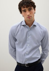 Camicia a maniche lunghe blu e bianca a righe, realizzata in tessuto intrecciato. Presenta un colletto, chiusura a bottoni e un design aderente. Abbinata a una cintura nera.