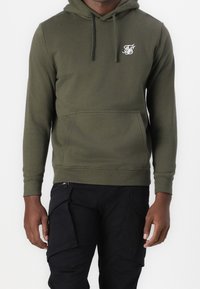Sudadera con capucha verde oliva hecha de una tela suave, con un bolsillo delantero y un pequeño logo blanco en el lado izquierdo del pecho. Pantalones cargo negros.