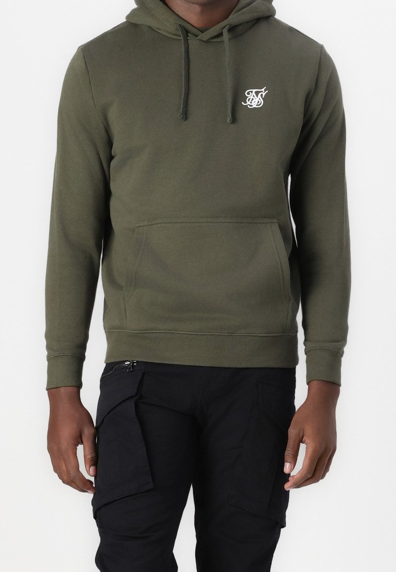 Sudadera con capucha verde oliva hecha de una tela suave, con un bolsillo delantero y un pequeño logo blanco en el lado izquierdo del pecho. Pantalones cargo negros.