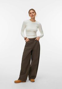 Femme portant un haut blanc à manches longues, un pantalon ample marron et des bottes beige, debout les mains dans les poches devant un fond uni.