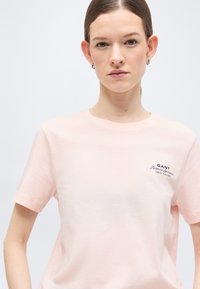 Γυναίκα με ίσια μαλλιά φοράει ροζ ανοιχτό t-shirt GANT με μικρό λογότυπο στο πάνω αριστερό μέρος του στήθους, στέκεται μπροστά σε απλό φωτεινό φόντο.