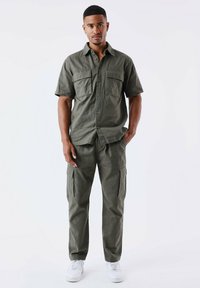 Maglietta verde oliva a maniche corte con due tasche frontali e pantaloni cargo abbinati con tasche laterali, abbinati a sneakers bianche.