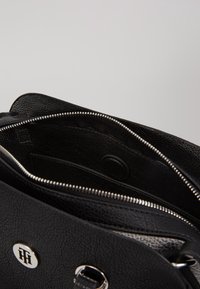 Bolso de mano de cuero negro texturizado con cierre de cremallera, herrajes en plata y un forro beig con estampado visible en el interior. Detalle de cierre redondo.