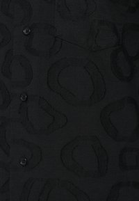 Fekete szövet nagy méretű, texturált absztrakt formákkal és fekete gombokkal, tűzött zsebfedéllel.