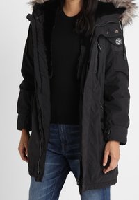 Sort parka-jakke med en hætte foret med kunstpels, lynlås- og knaplukning, sidelommer og en kviltet tekstur. Har et mærke på ærmet.