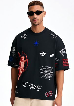 Jeune homme portant un T-shirt noir oversize avec un cupidon rouge, des yeux blancs, des phrases écrites et des motifs graphiques, associé à un pantalon beige et des lunettes de soleil noires.