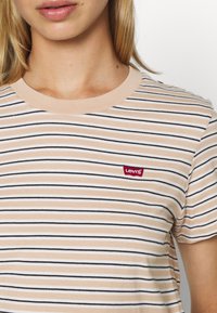 Ljusbeige randig t-shirt med horisontella svarta och vita ränder, en ribbad halsringning och en liten röd Levi's-logotyp på vänster bröst.