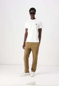 Tricou alb din bumbac cu un mic logo pe piept, combinat cu pantaloni tapered bej și adidași albi. Modelul poartă ochelari de soare.