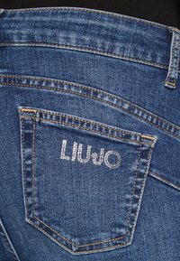 Jeans de mezclilla azul oscuro con un bolsillo trasero que presenta un logotipo "LIU JO" decorado con pequeños rhinestones y costuras amarillas contrastantes.