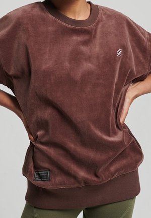 Osoba ubrana w luźną, krótką bluzkę z brązowego weluru z małym logo na piersi i naszywką przy dolnym brzegu.