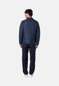 Giacca trapuntata blu con motivo a chevron, leggera, indossata con pantaloni neri e sneakers bianche, vista da dietro.