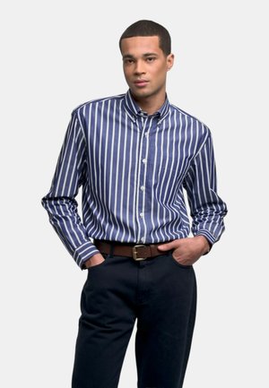 Uomo con capelli corti che indossa una camicia a bottoni a righe verticali blu e bianco, pantaloni blu navy e una cintura marrone, mani in tasca.