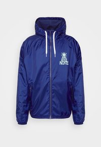 Mörkblå nylonvindjacka med dragkedja framtill, huva med dragsko, elastiska muddar och en ljusblå Nike-logotyp på bröstet.