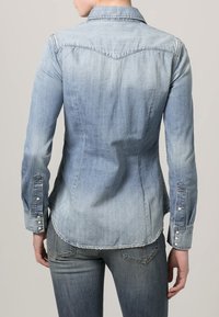 Chemise en denim bleu clair, avec une coupe ajustée, un col pointu et des poignets à boutons, présentant un subtil effet ombré sur le tissu.