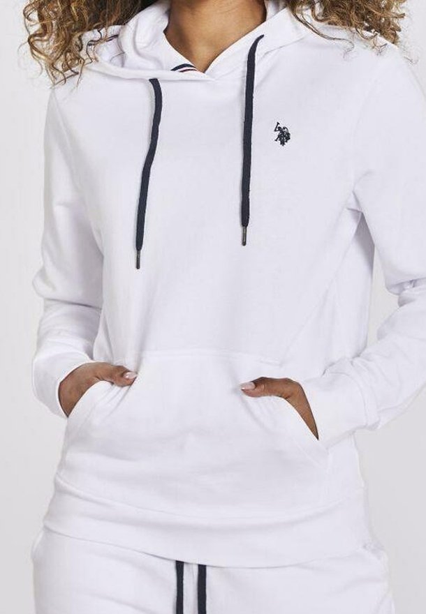 Personne portant un sweat à capuche blanc avec des cordons noirs et un petit logo noir brodé, les mains reposant dans la poche avant.