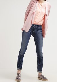 Cardigan rosa chiaro sopra una blusa pesca, abbinato a jeans skinny blu arrotolati e scarpe stringate metalliche. Texture morbide, vestibilità casual.