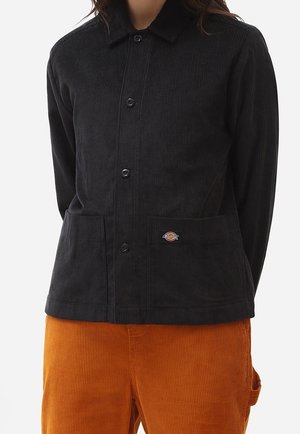 Person iført en sort manchetknap fløjlsjakke med forlommer og orange fløjlsbukser mod en ensfarvet baggrund.