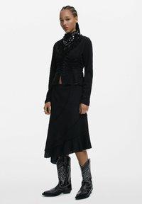 Top lungo a maniche nere con texture, dettagli arricciati, abbinato a una gonna nera fluente. Accentuato con una sciarpa a fantasia e stivali cowboy neri.