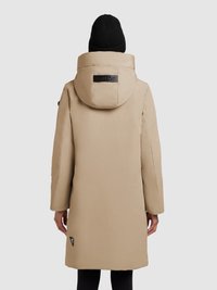 Lang beige polstret frakke med en stor hætte, sort logo-patch på ryggen og sidelommer. Varm, glat tekstur og løs pasform.