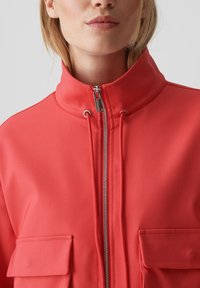 Rote Stoffjacke mit hohem Kragen, ausgestattet mit einem durchgehenden Reißverschluss und zwei vorderen Taschen. Glatte Textur, schlichtes Design ohne Muster.