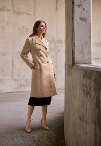 Beige dubbelzijdige jas met gouden knopen, zijzakken en een getailleerde pasvorm, gelaagd over een zwarte rok en nude hakken tegen een betonnen achtergrond.