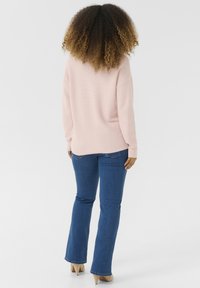 Svetlo roza, rebrasta pletenina s dolgimi rokavi in izrezanim robom, v kombinaciji z modrimi jeans hlačami v krogu in bež petami.