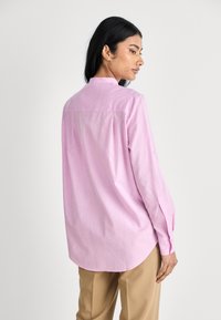 Camicia a maniche lunghe di un delicato rosa con colletto, dotata di una spalla posteriore dritta e polsini con bottoni, realizzata in tessuto leggero.