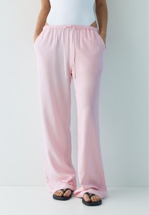 Osoba ubrana w luźne, jasnoróżowe spodnie z elastycznym paskiem i sznurkiem, stojąca z rękami w kieszeniach i czarnych klapkach.